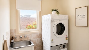 A laundry room with a washing machine, a dryer, a sink, and a window with a view of a brick wall and flowers. - Family-Friendly 2BR Villa with Private Courtyard (Malvern East)