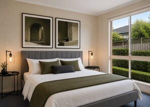 A bedroom with a large bed, two framed artworks above it, a nightstand with a lamp, and a window with a view of greenery. - Family-Friendly 2BR Villa with Private Courtyard (Malvern East)