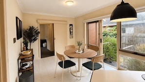 A dining area with a round table, chairs, and a pendant light. There is a window with a view of greenery outside. - Family-Friendly 2BR Villa with Private Courtyard (Malvern East)