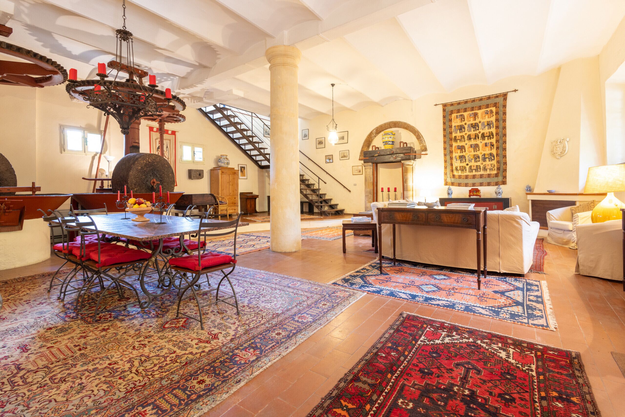 A spacious living area with a dining table, a chandelier, a staircase, and a large rug.