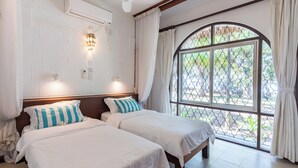 A hotel room with two beds, an air conditioning unit, a window with a view of greenery, and a ceiling fan. - Watano Beach House Spacious 7-bedroom house in awesome Diani perfect for groups (Diani)