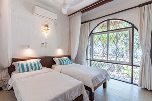 Ein Hotelzimmer mit zwei Betten, einer Klimaanlage, einem Fenster mit Blick auf Grünflächen und einem Deckenventilator.