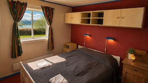 Un dormitorio con cama, ventana con cortinas, un armario de madera, una mesita de noche con lámpara, y vistas a la montaña.