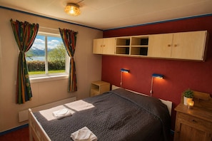 Ein Schlafzimmer mit Bett, einem Fenster mit Vorhängen, einem Holzschrank, einem Nachttisch mit Lampe und Blick auf Berge.