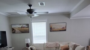 A living room with a sectional sofa, a TV, a ceiling fan, and artwork on the walls. - Cozy 2-bedroom house nestled among the trees and surrounded by open space. (Eastman)