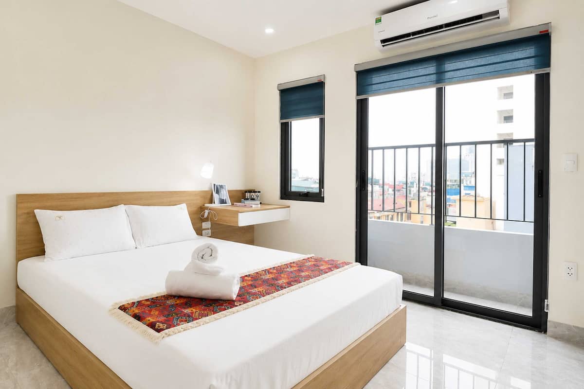 A hotel room with a bed, bedside table, window, and air conditioning unit.