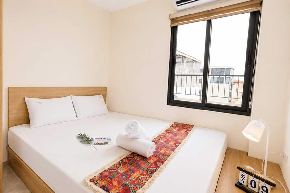 A hotel room with a bed, a window with a view, a bedside lamp, and a wall clock.
