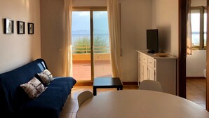 Ein Zimmer mit einer Couch, einem Fernseher auf einem Schrank, einem Tisch und Meerblick durch eine Schiebetür.