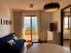 Een kamer met een bank, een televisie op een kast, een tafel en uitzicht op de zee door een schuifdeur.