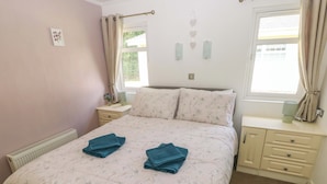 Un dormitorio con colcha estampada, dos toallas sobre la cama, una mesita de noche con lámpara y una ventana con cortinas.