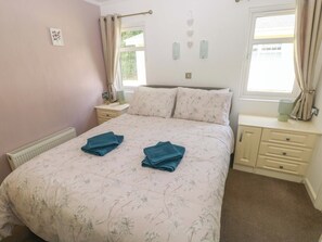 A bedroom with a floral bedspread, two towels on the bed, a nightstand with a lamp, and a window with curtains.
