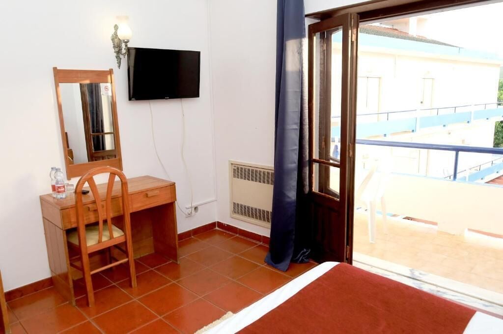A room with a wooden desk, a chair, a television mounted on the wall, and a window with a view of a building outside.