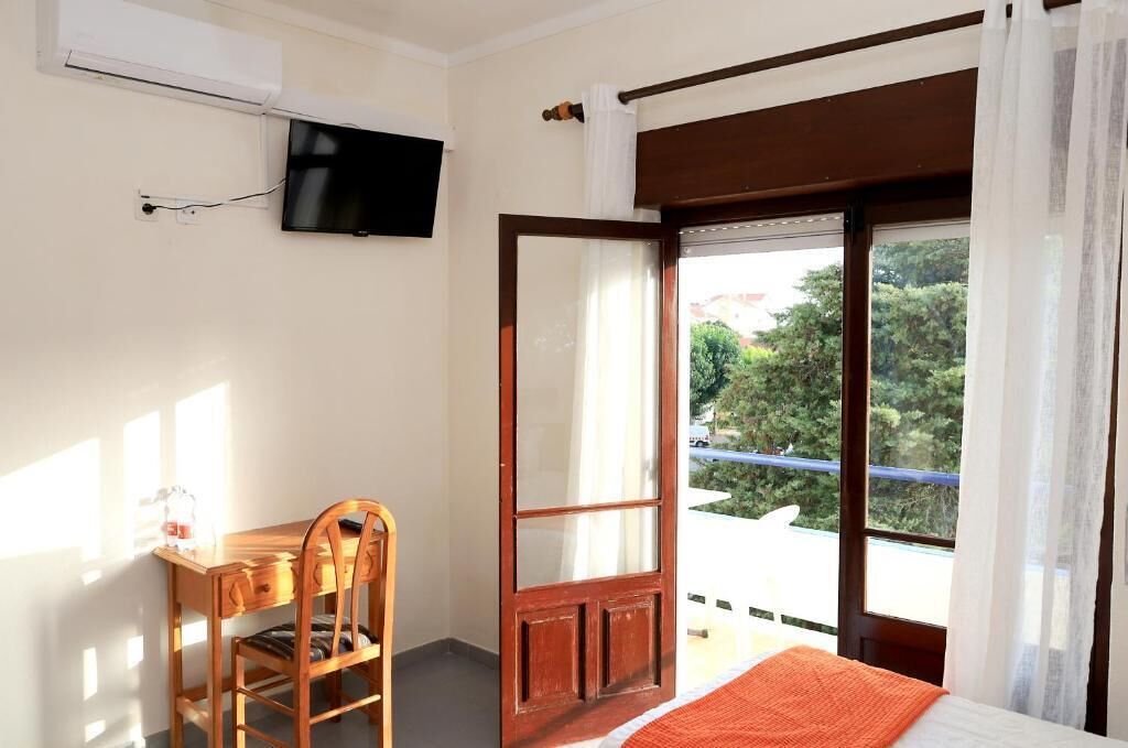 A room with a bed, a desk, a chair, a TV mounted on the wall, and a window with a view of greenery and a building.