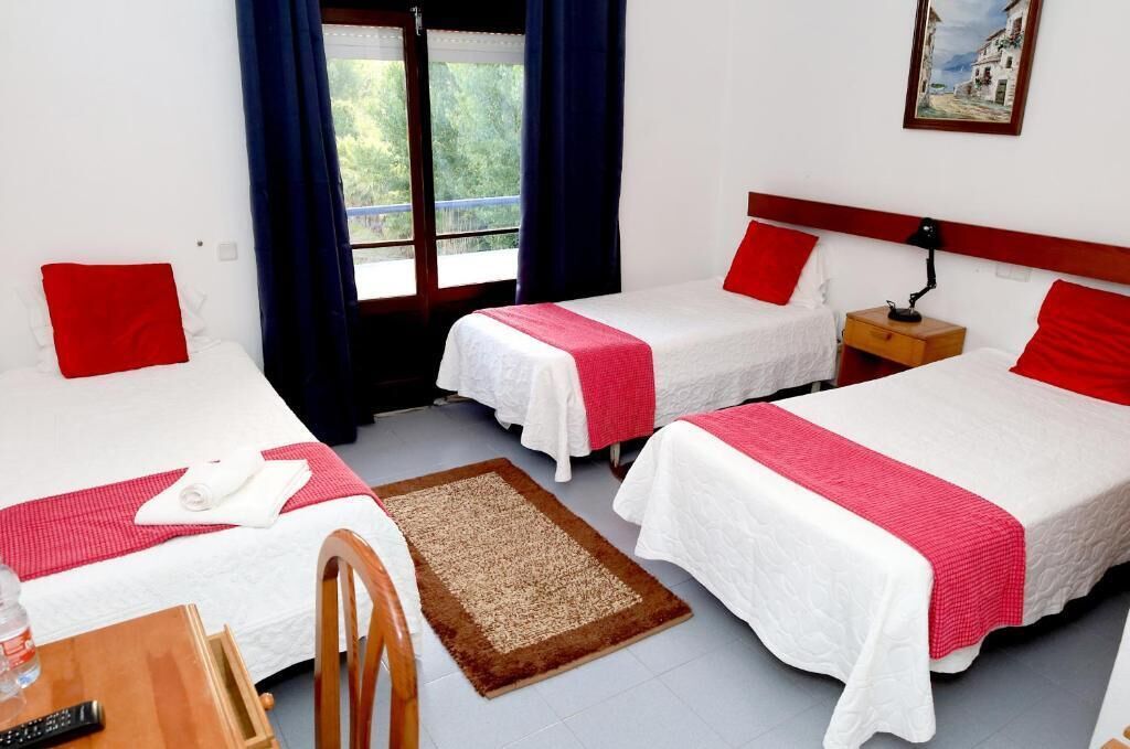 A hotel room with two beds, a wooden nightstand, a chair, a small table, and a window with curtains.