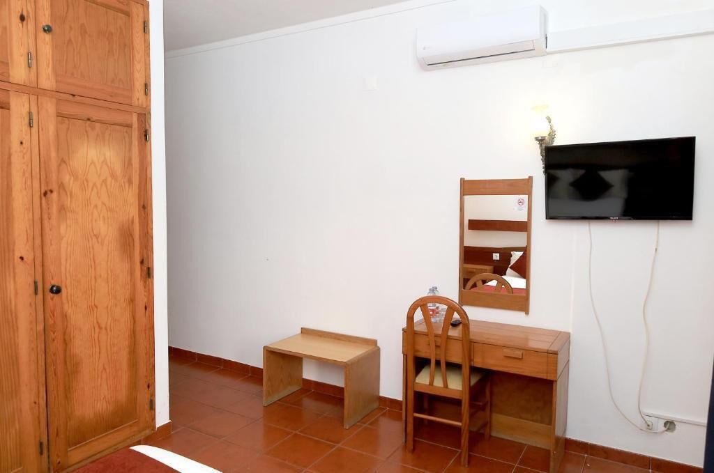 A room with a wooden wardrobe, a desk with a chair, a small bench, a television mounted on the wall, and an air conditioning unit.