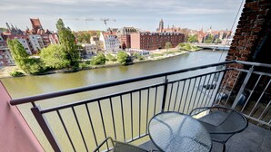 A balcony with two chairs and a table overlooking a river with buildings in the background. - Live & Travel Apartments Waterlane  (Gdańsk)