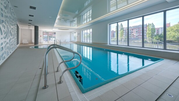 An indoor swimming pool with clear glass walls, a tiled floor, and a metal pool ladder. - Live & Travel Apartments Waterlane  (Gdańsk)