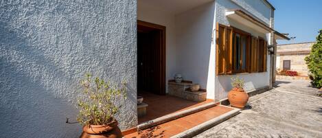 A building with a white exterior, a wooden door, and a small porch area with potted plants.