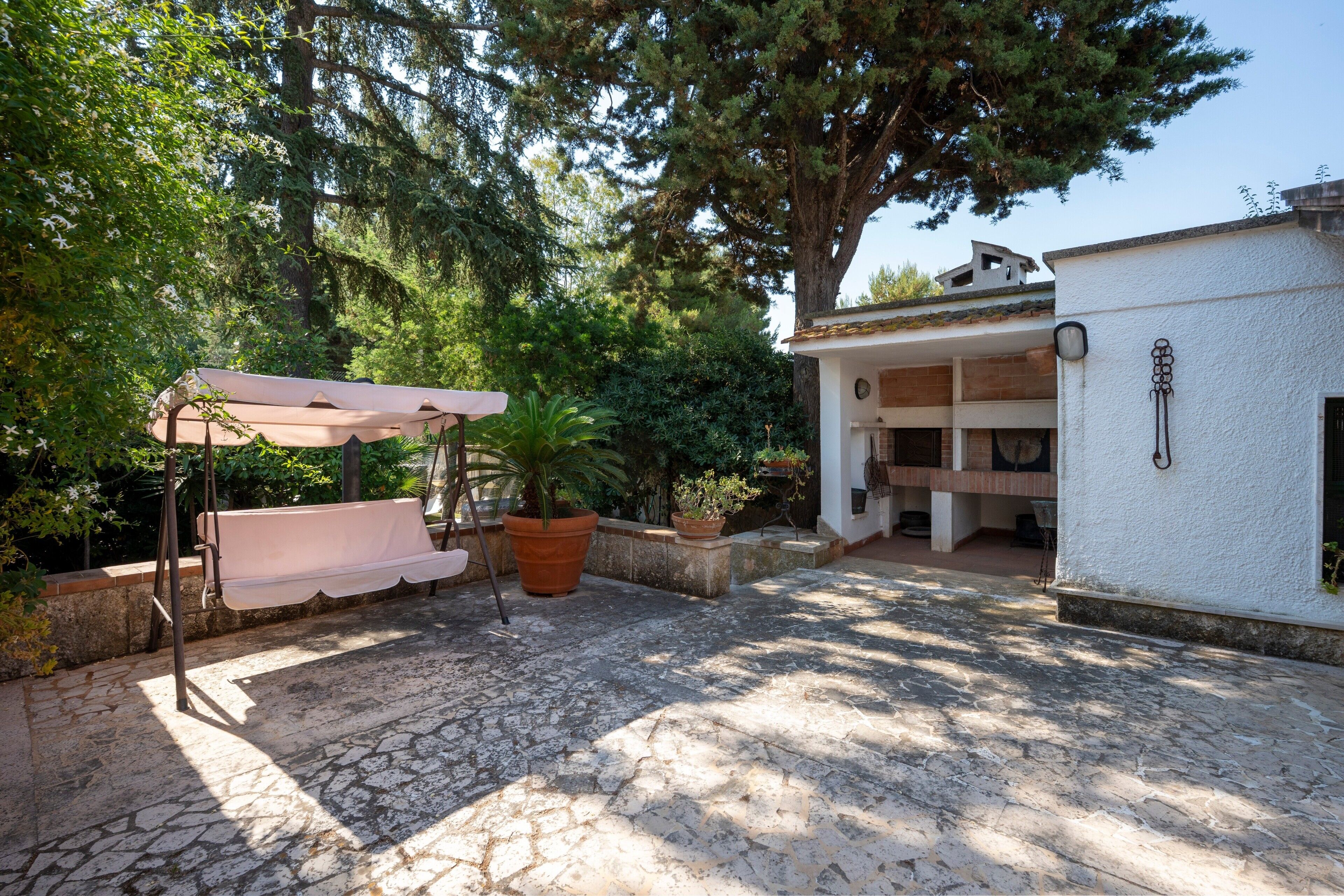 A stone patio with a bench and potted plants, a white building with a balcony, and a large tree providing shade.