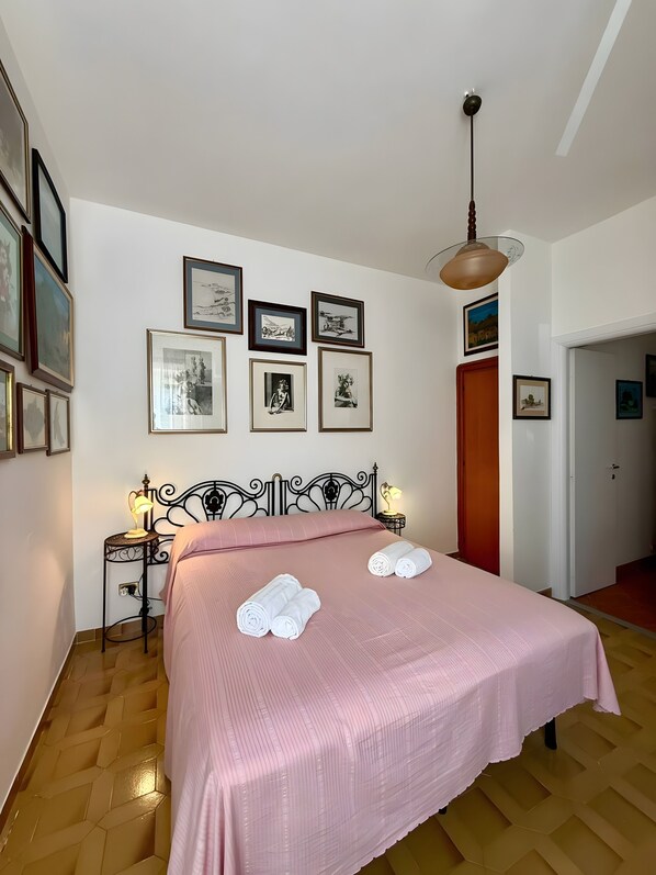Ein ordentlich bezogenes Bett mit einer rosa Tagesdecke, zwei aufgerollte Handtücher und eine Wand mit gerahmten Bildern.