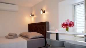 Une chambre à coucher comprenant un lit, un bureau, deux chaises, une fenêtre avec des stores et un climatiseur fixé au mur.