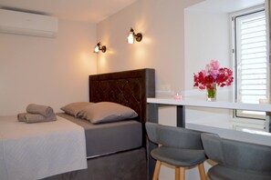 A bedroom with a bed, a desk, two chairs, a window with blinds, and a wall-mounted air conditioner. - Groundfloor Apartment Landula (Preko)