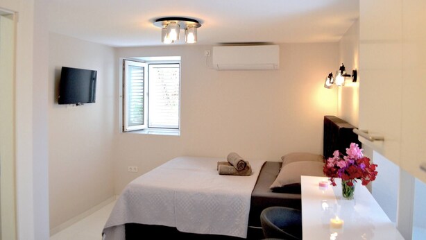 Une chambre d’hôtel comprenant un lit, un bureau, une télévision, un climatiseur, une fenêtre avec des stores et un vase avec des fleurs.