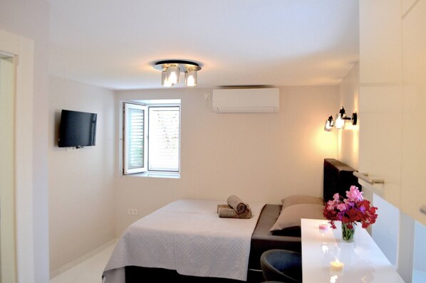 A hotel room with a bed, a desk, a TV, an air conditioner, a window with blinds, and a vase with flowers. - Groundfloor Apartment Landula (Preko)