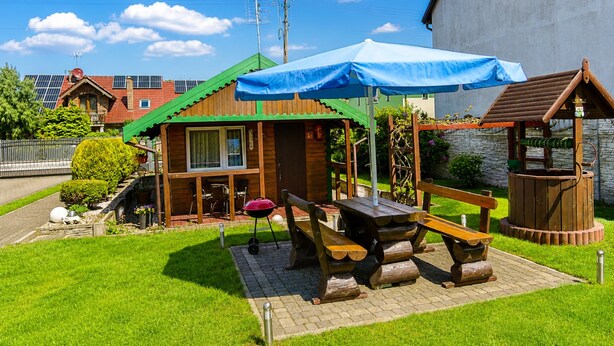 Uma cabana de madeira com mesa de piquenique e bancos, churrasqueira e toldo azul.