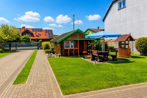 Ein Wohnhaus mit befestigter Auffahrt, ein Holzhaus mit Gründach, einem Grillplatz und einem kleinen Pavillon mit blauer Markise.
