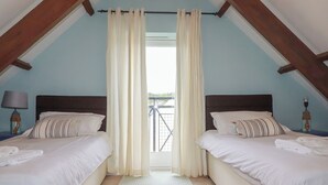Ein Schlafzimmer mit zwei Betten, einem Fenster mit Vorhängen, einem Nachttisch mit Lampe und Blick auf einen Balkon.
