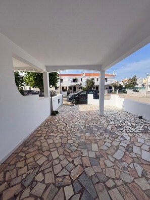 A covered outdoor area with a stone-tiled floor, white walls, and a view of a building with a red roof. - Villa Praia (Altura)