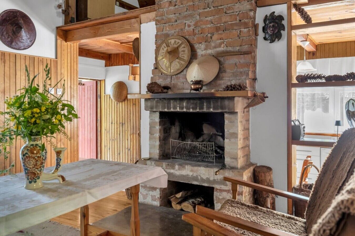 A rustic interior with a brick fireplace, wooden beams, and a table with a vase of flowers.