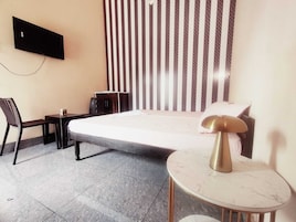 Quarto de hotel com cama, escrivaninha, cadeira, mesinha, televisão na parede e cortina estampada.