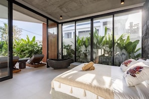 A modern bedroom with a large bed, a bathtub, and a view of greenery through large windows. - Villa Karma | 2BR pool villa in Seseh (Mengwi)