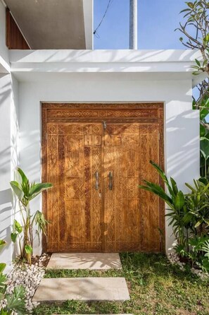 A wooden door with intricate patterns, a concrete pathway, and surrounding greenery. - Villa Karma | 2BR pool villa in Seseh (Mengwi)