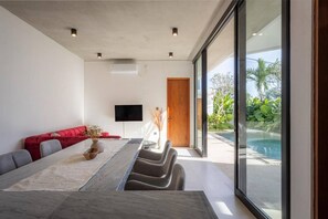 A modern living room with a long dining table, red sofa, and a TV on the wall. - Villa Karma | 2BR pool villa in Seseh (Mengwi)