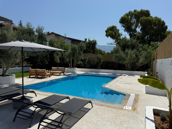 Une piscine entourée de verdure et de bâtiments, avec des chaises longues et un parasol.