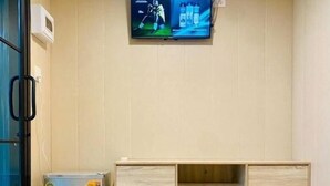 Quarto com piso de madeira, televisão fixada na parede, pequena geladeira e armário de madeira.