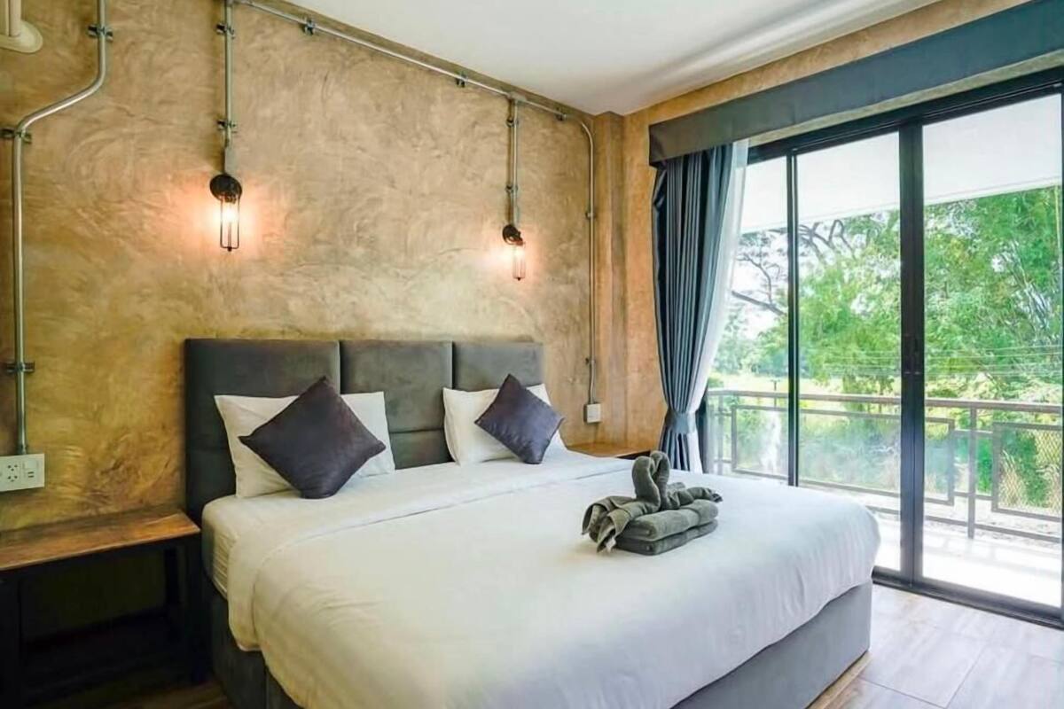 A modern bedroom with a large bed, a nightstand, and a view of greenery through the window.