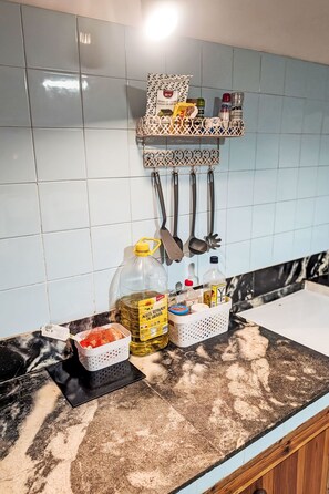 Et køkken med flisebeklædt bagvæg, en ophængt grydeholder, en flaske madolie, en kurv og en sukkerdåse.