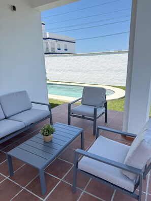 A patio with a sofa, chairs, and a table, overlooking a pool and a white building.