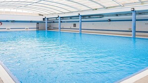 Piscina interna com teto de vidro curvo, água cristalina e azulejos azuis na borda.
