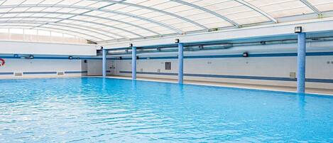 An indoor swimming pool with a curved glass ceiling, clear water, and blue poolside tiles.