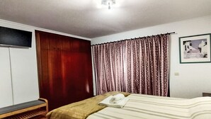 Quarto com cama, um banco de madeira, guarda-roupa, cortinas estampadas e um quadro na parede.