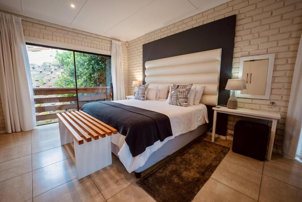 Une chambre à coucher avec un lit, un banc, une table de chevet, une lampe et une vue sur la verdure à l’extérieur.