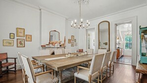 Uma sala de jantar com uma grande mesa de madeira, cadeiras estofadas, um candelabro e quadros nas paredes.
