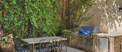 An outdoor patio with a wooden table and chairs, a barbecue grill, and a brick wall covered in ivy.