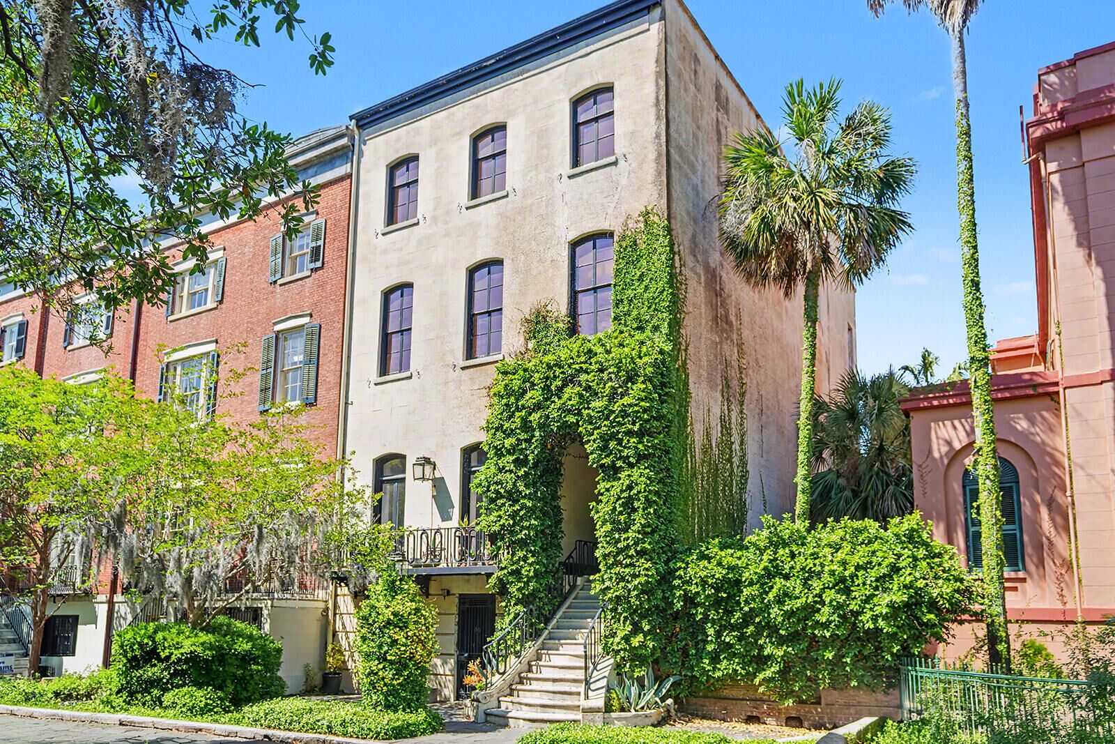 A multi-story building with a brick facade, a staircase leading to the entrance, and lush greenery around the property.