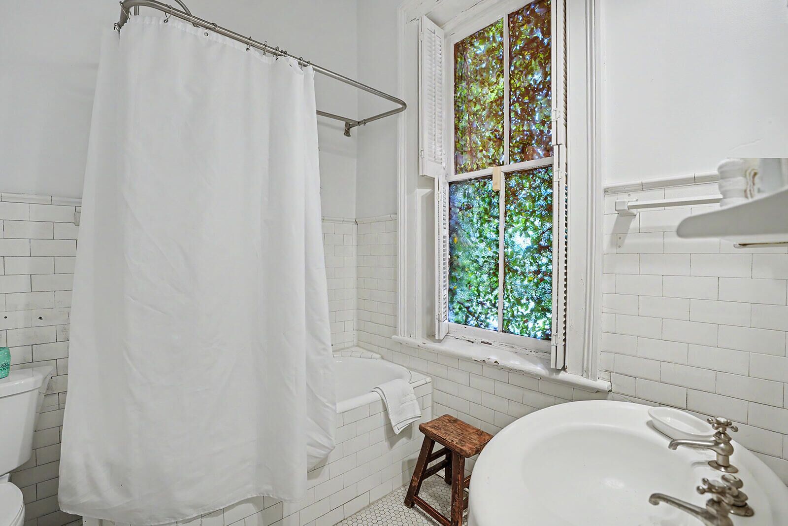 Une salle de bain avec une baignoire blanche, un rideau de douche, une fenêtre avec des vitraux et un tabouret en bois.